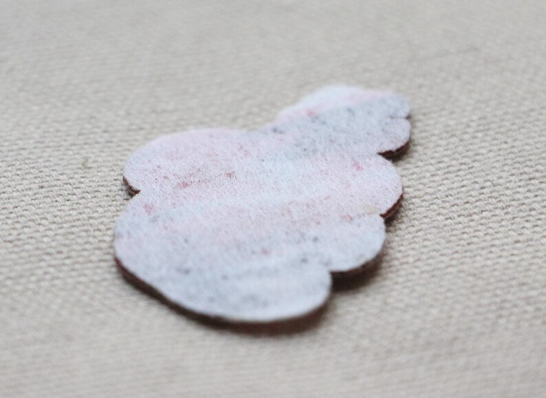 May include: A small, light-coloured felt appliqu&eacute; in a floral shape. The felt is white with a subtle pattern of pink and blue. The edges are a dark brown colour. The appliqu&eacute; is likely for crafting or embellishment.