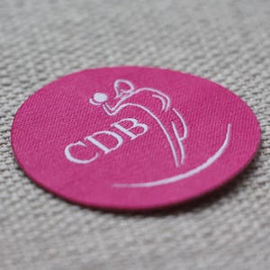 May include: A circular, cerise-coloured patch featuring white embroidered text and a stylised figure. The text reads "CDB" and is accompanied by a curved line and a figure in motion, possibly playing a sport.