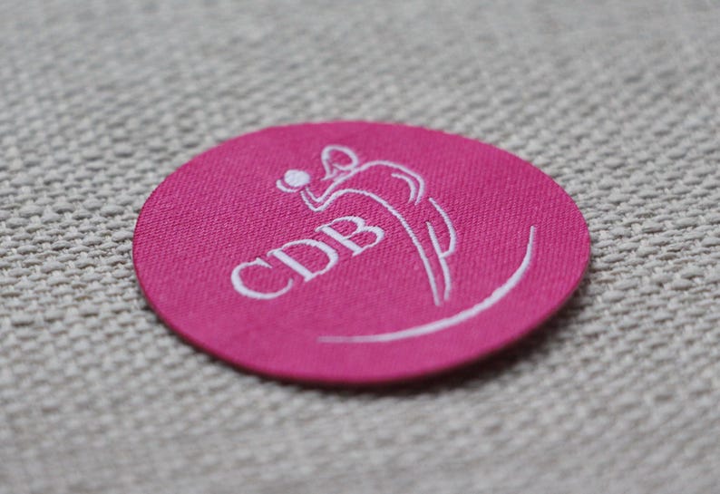 May include: A circular, cerise-coloured patch featuring white embroidered text and a stylised figure. The text reads "CDB" and is accompanied by a curved line and a figure in motion, possibly playing a sport.