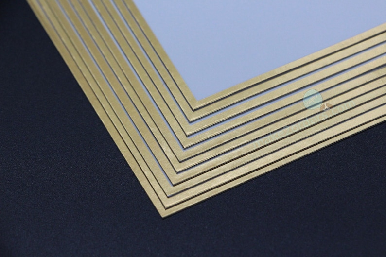 May include: Close-up of a gold-coloured, layered frame with a geometric design. The frame is made of multiple thin, parallel bars, creating a textured surface. The background is a dark blue-grey colour.