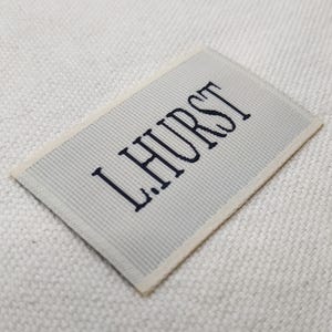 May include: A rectangular label with the text "L.HURST" embroidered in dark blue on a light gray striped background. The label has a cream-colored border and is set against a textured white surface.