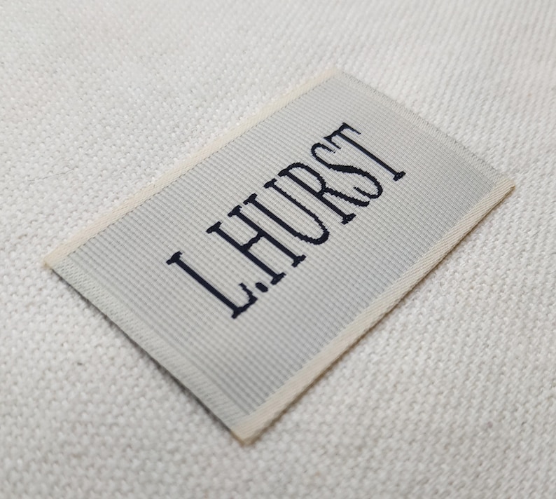 May include: A rectangular label with the text "L.HURST" embroidered in dark blue on a light gray striped background. The label has a cream-colored border and is set against a textured white surface.