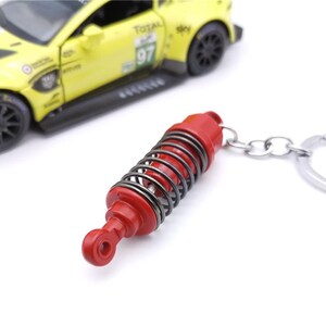May include: A red and silver keychain featuring a miniature shock absorber. The keychain has a silver spring and a red body. A yellow toy race car is in the background.