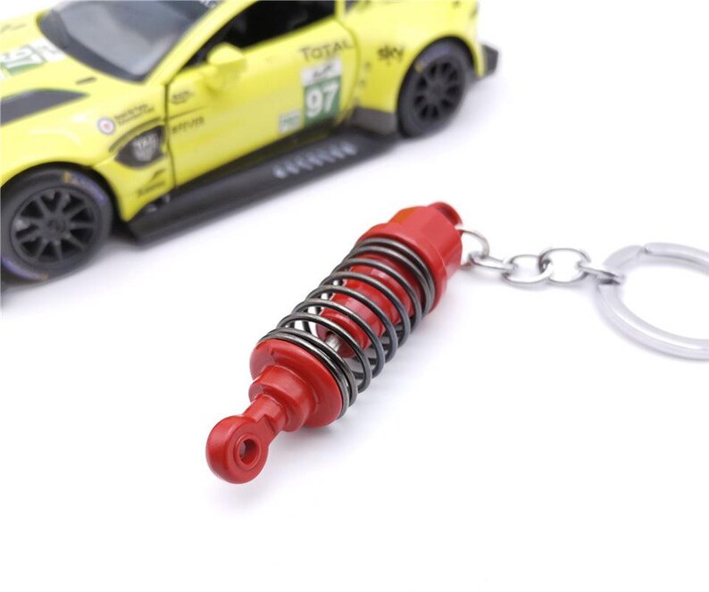May include: A red and silver keychain featuring a miniature shock absorber. The keychain has a silver spring and a red body. A yellow toy race car is in the background.