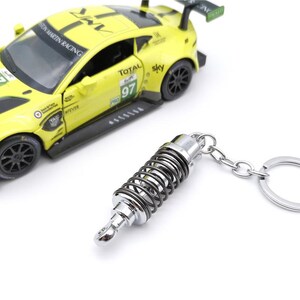 May include: A yellow race car model with black accents and the number 97 is next to a silver and black shock absorber keychain. The keychain has a silver ring and chain.