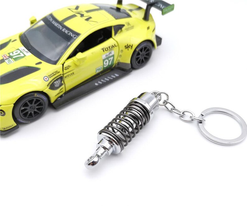 May include: A yellow race car model with black accents and the number 97 is next to a silver and black shock absorber keychain. The keychain has a silver ring and chain.