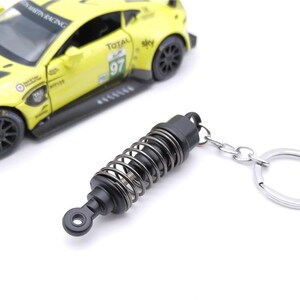 May include: A black and silver keychain featuring a miniature shock absorber design. The keychain has a silver key ring and a spring coil detail. A yellow toy race car is in the background.
