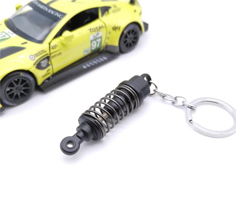 May include: A black and silver keychain featuring a miniature shock absorber design. The keychain has a silver key ring and a spring coil detail. A yellow toy race car is in the background.
