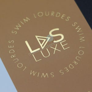 May include: A brown card with gold lettering, featuring the brand name "LVS LUXE" and the text "SWIM LOURDES" in a circular pattern. The design is minimalist and elegant, suggesting a high-end product.