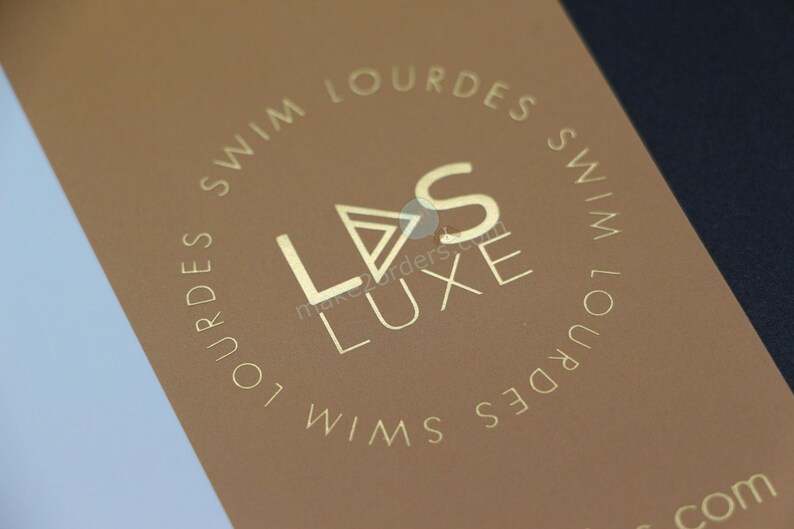 May include: A brown card with gold lettering, featuring the brand name "LVS LUXE" and the text "SWIM LOURDES" in a circular pattern. The design is minimalist and elegant, suggesting a high-end product.