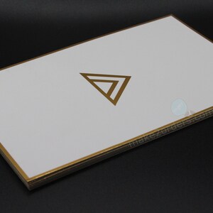 May include: A stack of white cards with gold edges and a gold geometric logo. The cards are rectangular and have a clean, minimalist design. The gold accents provide a touch of elegance. The cards are likely used for business or promotional purposes.