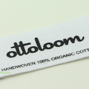 May include: A white rectangular fabric label with the black text "ottoloom" in a cursive font. Below, the text reads "HANDWOVEN 100% ORGANIC COTTON". The label is on a light background.
