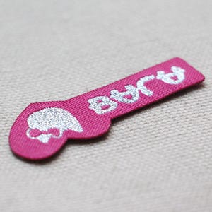 May include: A pink fabric label with the word "BAJA" in white, stylized text. The label has a rounded end with a skull and bow design in white. The label is likely for clothing or accessories.