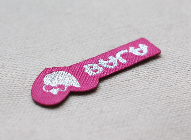 May include: A pink fabric label with the word "BAJA" in white, stylized text. The label has a rounded end with a skull and bow design in white. The label is likely for clothing or accessories.
