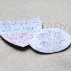 May include: Two small, textured fabric patches in a light colour palette. One is a rounded shape, and the other is a half-circle. The patches have a speckled appearance with hints of blue, pink, and grey.