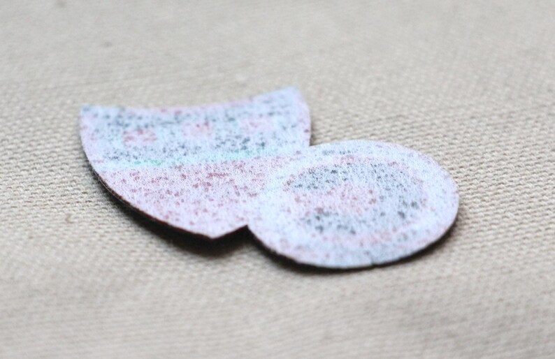 May include: Two small, textured fabric patches in a light colour palette. One is a rounded shape, and the other is a half-circle. The patches have a speckled appearance with hints of blue, pink, and grey.