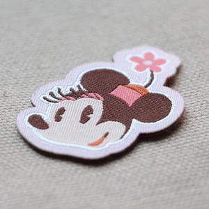 May include: A fabric patch depicting a cartoon mouse head with brown ears, a pink bow, and a flower. The patch has a light pink outline and is set against a neutral background.
