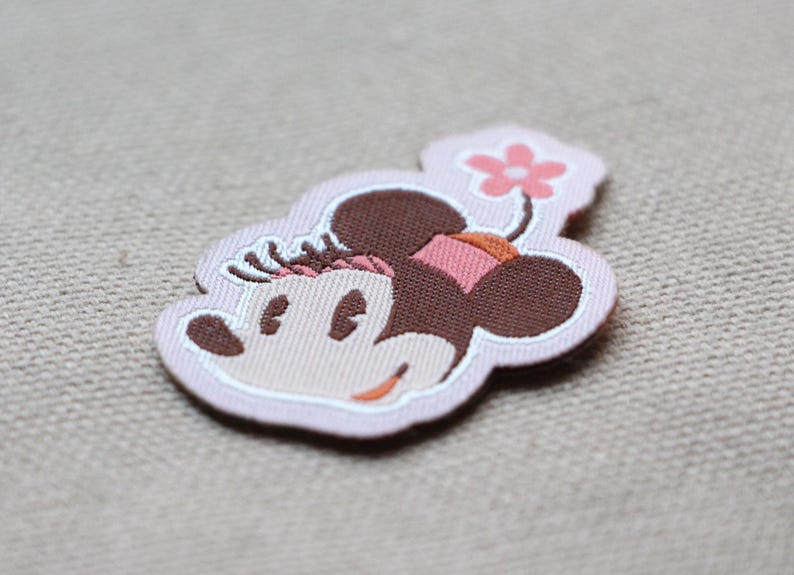 May include: A fabric patch depicting a cartoon mouse head with brown ears, a pink bow, and a flower. The patch has a light pink outline and is set against a neutral background.