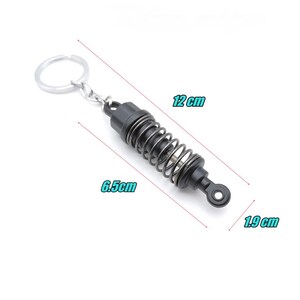 May include: Black and silver keychain featuring a miniature shock absorber design. The keychain measures 4.7 inches long, 2.6 inches wide, and 0.7 inches thick. The spring is silver, and the rest of the keychain is black.