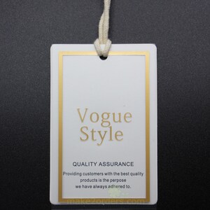 May include: A white rectangular tag with a gold border and a cream-colored string. The tag features the words "Vogue Style" in gold lettering, along with additional text about quality assurance. The background is dark.