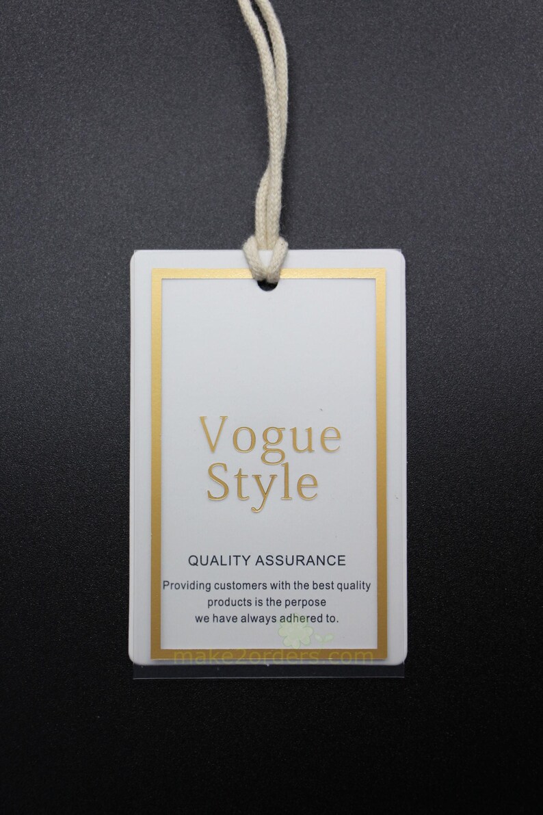 May include: A white rectangular tag with a gold border and a cream-colored string. The tag features the words "Vogue Style" in gold lettering, along with additional text about quality assurance. The background is dark.