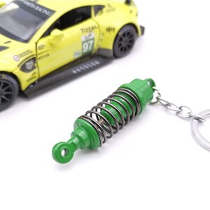 May include: A green and silver metal keychain shaped like a car suspension spring. The keychain is next to a yellow toy race car with black wheels. The car has the number 97 on the side.