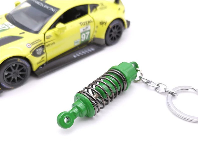 May include: A green and silver metal keychain shaped like a car suspension spring. The keychain is next to a yellow toy race car with black wheels. The car has the number 97 on the side.