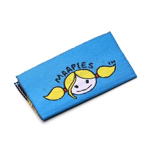 以下が含まれることがあります： 「MAAPIES」の文字と、金髪のおさげ髪の女の子の漫画が描かれた青い長方形のラベル。ラベルは白い背景にあります。ラベルは布製です。