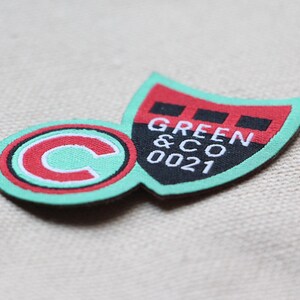 May include: A colourful fabric patch featuring a shield and a circular emblem. The shield is red and black with the text "GREEN & CO 0021" in white. The circle has a red border and a white "C" on a mint green background.
