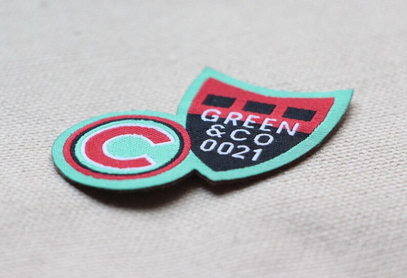May include: A colourful fabric patch featuring a shield and a circular emblem. The shield is red and black with the text "GREEN & CO 0021" in white. The circle has a red border and a white "C" on a mint green background.