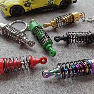 May include: A collection of colorful shock absorber keychains. The keychains feature a spring design with various colors, including silver, green, gold, black, red, and iridescent. A yellow toy car is in the background.