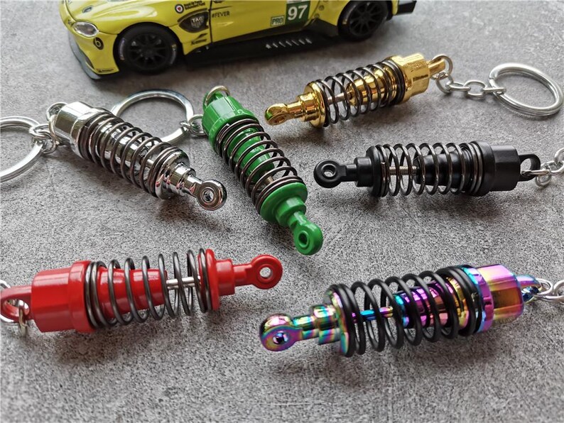 May include: A collection of colorful shock absorber keychains. The keychains feature a spring design with various colors, including silver, green, gold, black, red, and iridescent. A yellow toy car is in the background.