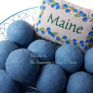 May include: A blue wire basket filled with blue bath bombs. A small pillow with the word "Maine" embroidered on it, surrounded by a blueberry design, rests in the basket. Text on the image reads "Welcome to.. The Soapmaker's Shop of Maine."