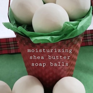 May include: A red bucket filled with white shea butter soap balls, resting on a green surface. The bucket has a handle and is lined with green tissue paper. The text on the bucket reads "moisturizing shea butter soap balls."