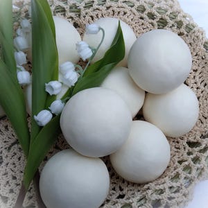 May include: A collection of white, round objects, possibly food items, arranged in a woven basket. Green leaves and delicate white lily of the valley flowers are interspersed among the objects, adding a natural touch to the composition.