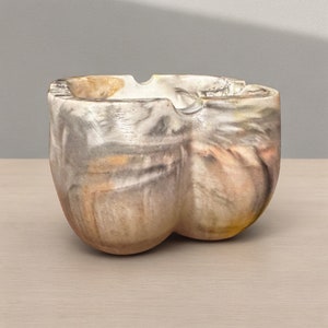 Puede incluir: Un cenicero de cerámica con un diseño abstracto único en tonos grises, marrones y amarillos. El cenicero tiene una forma redondeada, orgánica y un acabado liso y brillante.