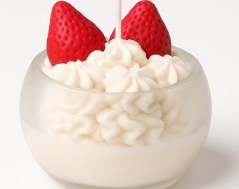 May include: A dessert-themed candle in a clear glass container. The candle features a white, whipped cream-like design topped with two red strawberry-shaped elements. A white wick extends from the center. The candle is designed to resemble a dessert.