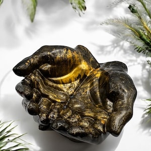 May include: A golden tiger's eye stone carved into the shape of two hands cupped together. The stone has a black and gold color pattern.