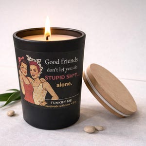 May include: A black candle with a lit wick and a wooden lid. The candle's label features a vintage illustration of two women and the text "Good friends don't let you do STUPID SH*T... alone."