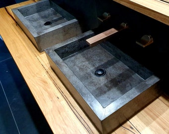 Op de afbeelding: Twee rechthoekige betonnen wastafels met een donkergrijze afwerking zijn gemonteerd op een houten aanrechtblad. Het aanrechtblad is gemaakt van lichtbruin hout met donkerbruine strepen. De wastafels zijn elk uitgerust met een enkele kraan met een koperen afwerking.