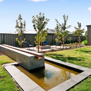 May include: A modern garden with a concrete water feature and a fire pit. The water feature is surrounded by a grey stone patio and a lush green lawn. There are several trees and shrubs in the background.
