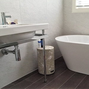May include: A white bathroom sink with a chrome tap and a white countertop. A white bath is in the background. A small round stool with a white and grey speckled surface is in the foreground.