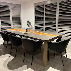 May include: A large dining table with a concrete top and a live edge wood inlay. The table has a gold metal base and is surrounded by six black chairs with gray fabric seats.