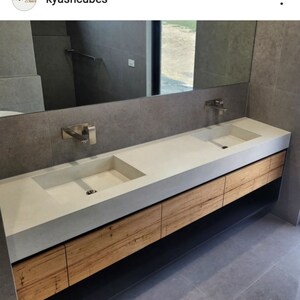May include: A double vanity featuring a long, light gray concrete countertop with two integrated rectangular sinks. The base consists of horizontal light brown wood grain drawers and a black lower shelf, paired with two wall-mounted silver faucets.
