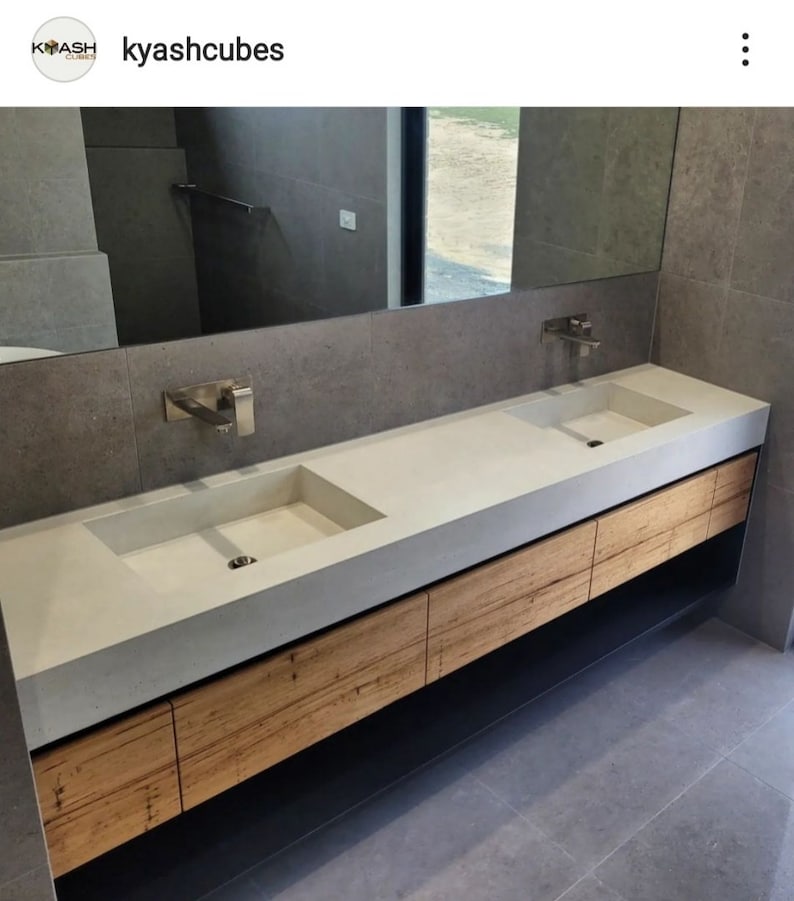 May include: A double vanity featuring a long, light gray concrete countertop with two integrated rectangular sinks. The base consists of horizontal light brown wood grain drawers and a black lower shelf, paired with two wall-mounted silver faucets.
