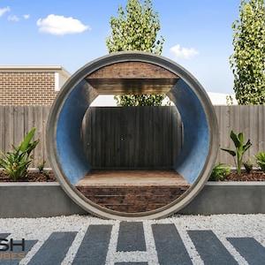May include: A large concrete pipe, cut in half, is used as a unique outdoor structure. The pipe is painted blue on the inside and has a wooden platform inside. The pipe is surrounded by gravel and plants.