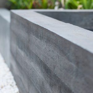 May include: A close-up of a gray concrete planter with a textured surface. The planter is made of stacked concrete blocks.