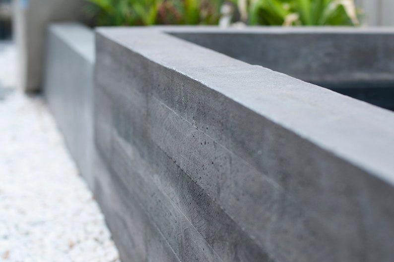 May include: A close-up of a gray concrete planter with a textured surface. The planter is made of stacked concrete blocks.
