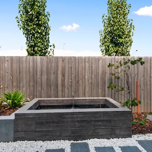 May include: A rectangular concrete water feature with a small fountain in a backyard setting. The water feature is surrounded by white gravel and a wooden fence.