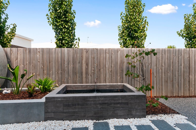 May include: A rectangular concrete water feature with a small fountain in a backyard setting. The water feature is surrounded by white gravel and a wooden fence.